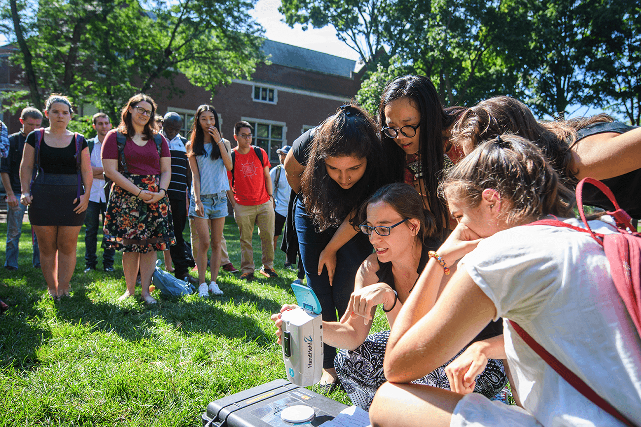 Image of students outdoors, crowded around female professor as she shows them a piece of remote sensing equipment.