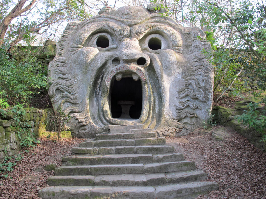 Steps leading up to a gray stone cave surrounded by greenery. The cave has a face carved into it with a large mouth over the entry way, eyes, and a nose. 