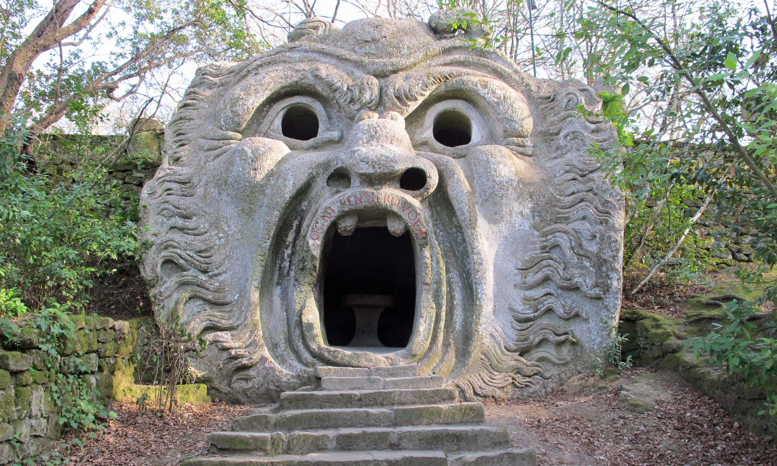 Steps leading up to a gray stone cave surrounded by greenery. The cave has a face carved into it with a large mouth over the entry way, eyes, and a nose.