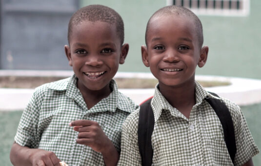 Two school children smiling at the camera, wearing uniforms and backpacks