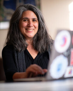 Director of Sustainability and Social Justice Laurie Ross working at a computer, smiles at the viewer