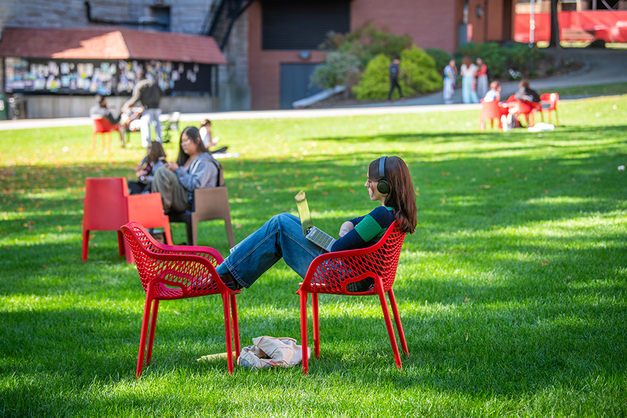 student listening to laptop in chair on academic campus grounds