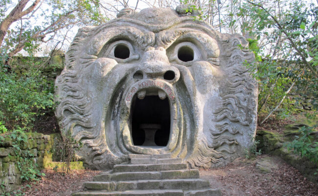 Steps leading up to a gray stone cave surrounded by greenery. The cave has a face carved into it with a large mouth over the entry way, eyes, and a nose.