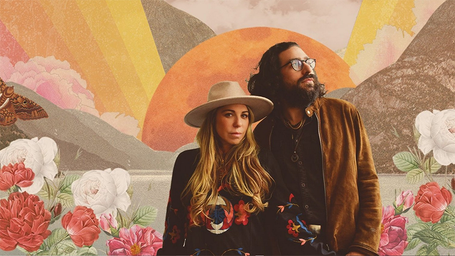 Nefesh Mountain band members Eric Lindberg and Doni Zasloff stand in front of a colorful mural with white and red flowers and an orange sunrise on the horizon.
