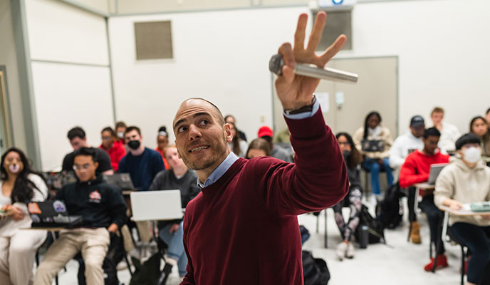 A faculty member holding a microphone makes reference to the presentation space while lecturing to a classroom of students