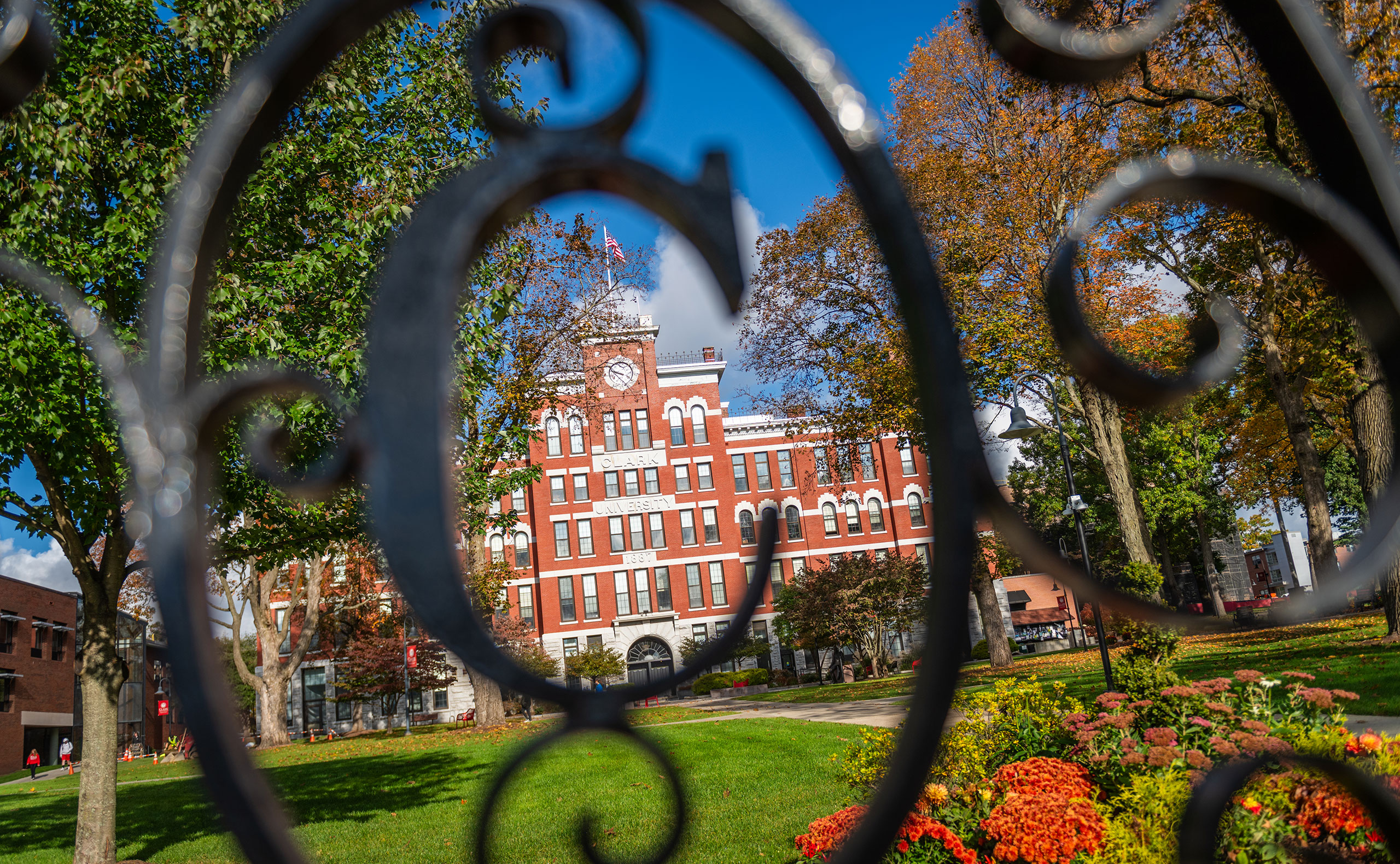 The letter 'C' on a wrought iron fence with Jonas Clark Hall in the background, fall 2023
